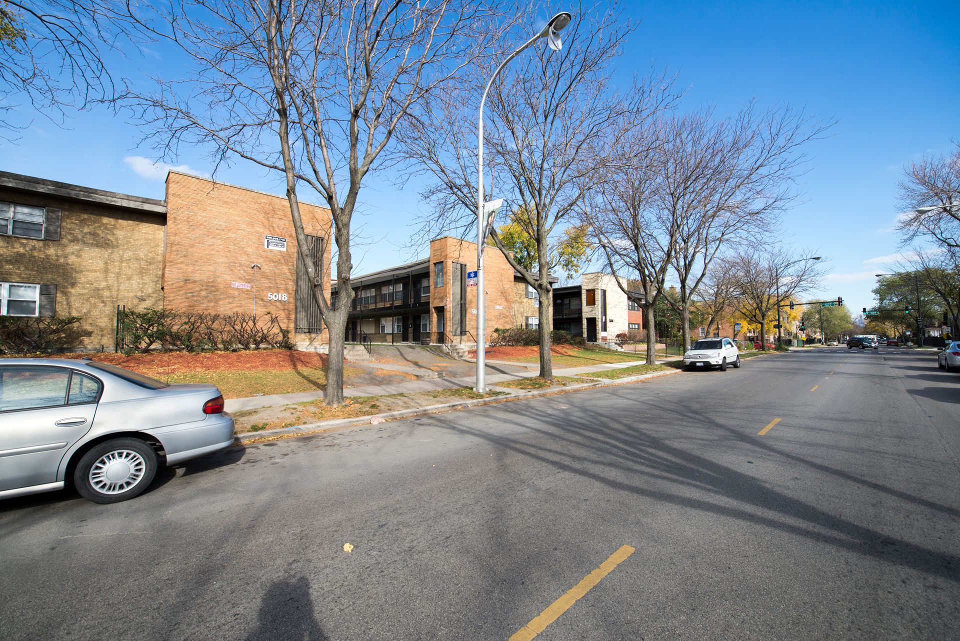 Apartment buildings line a street on a sunny day. A silver car is parked on the side.