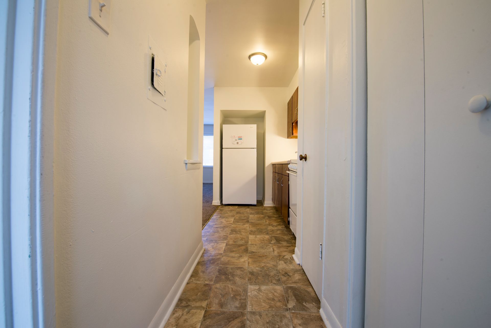 Narrow hallway with a linoleum floor, leading to a kitchen with a refrigerator visible.
