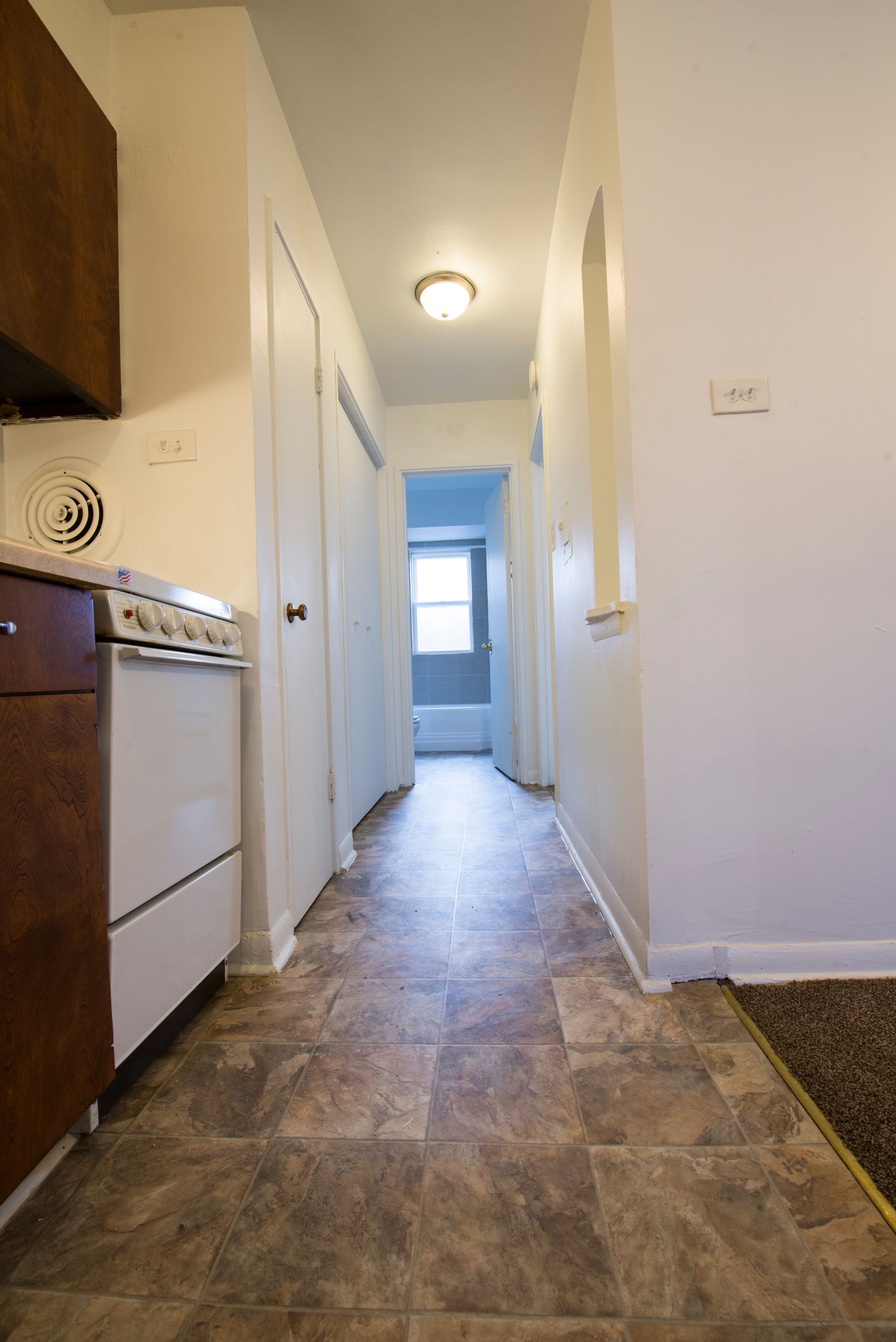 Narrow hallway with brown tiled floor, leading to a window. White walls, doors, and a light fixture.