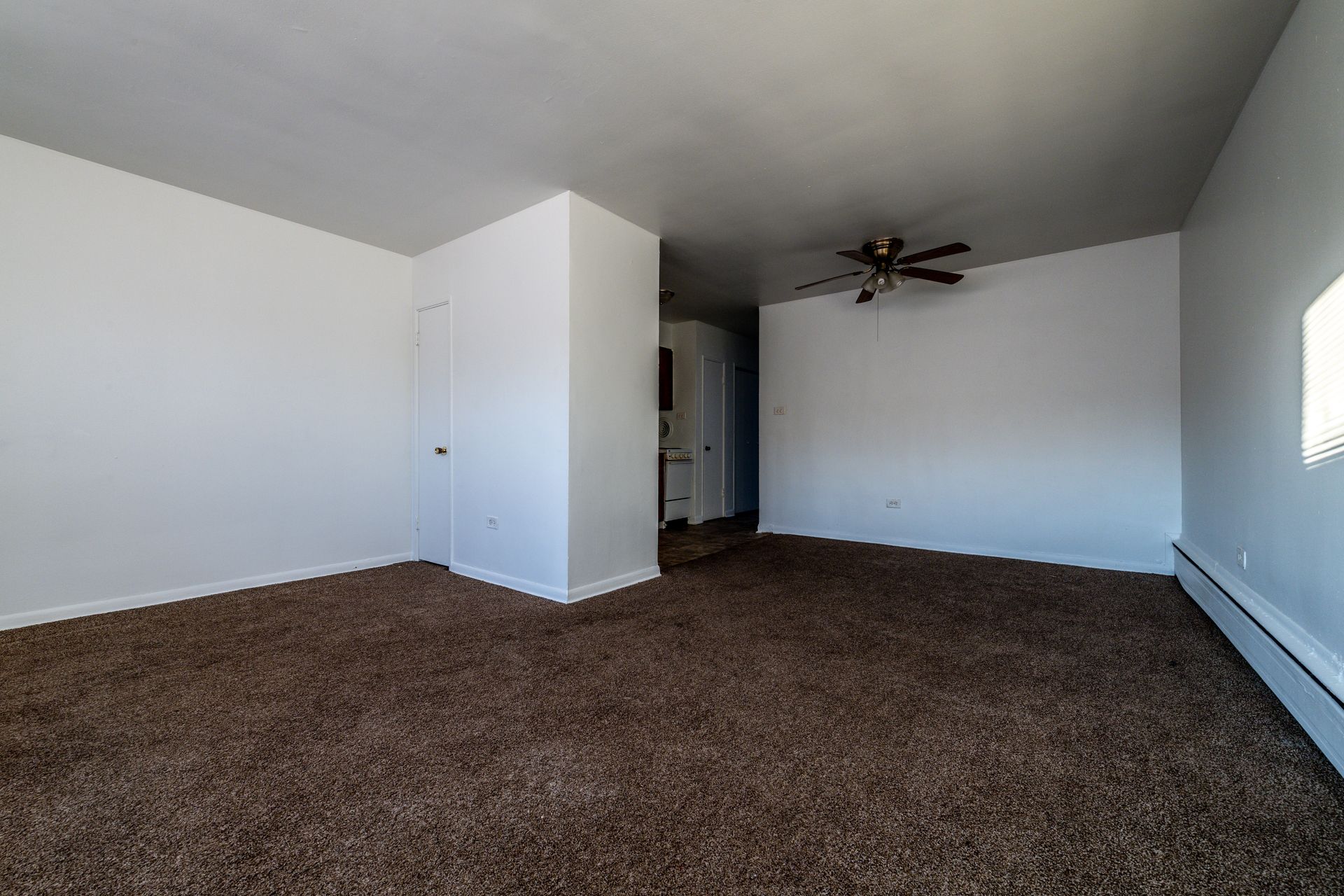 Empty room with brown carpet, white walls, and a ceiling fan. A door and radiator are visible.