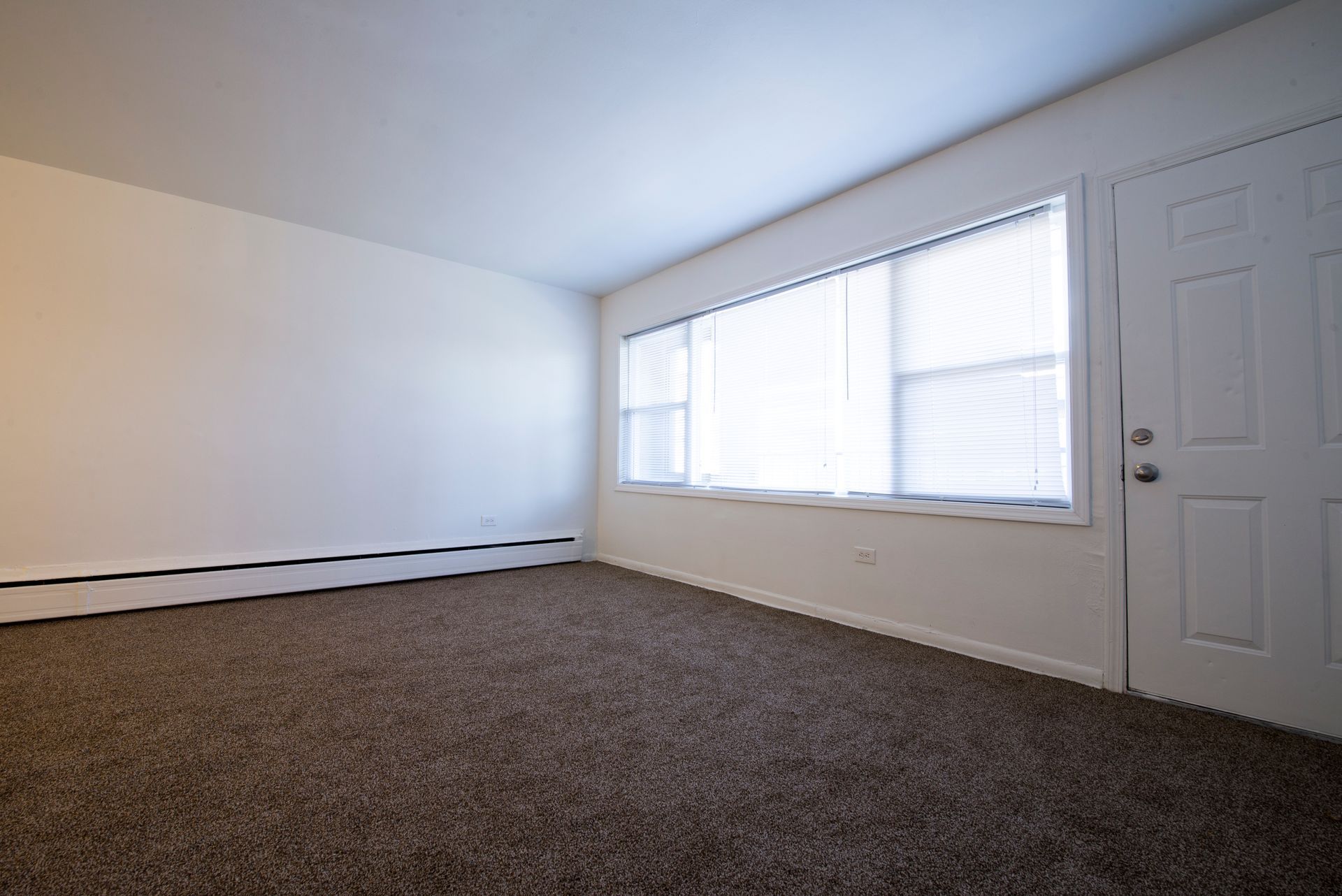 Empty room with brown carpet, white walls, a window with blinds, and a door.