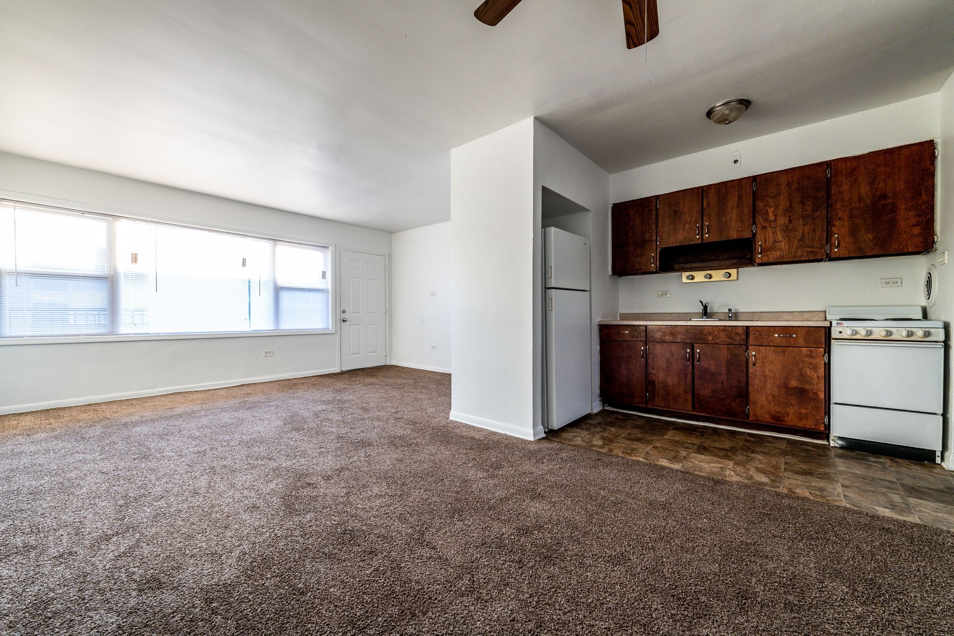 An empty apartment interior with carpet, a kitchen area with dark cabinets, and a large window.