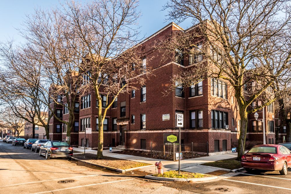 Brick apartment building on a street corner, cars parked along the street, trees with bare branches, sunny day.