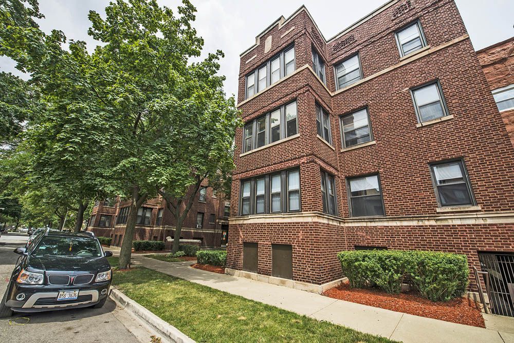 Red brick apartment building with a parked car and a tree in front.