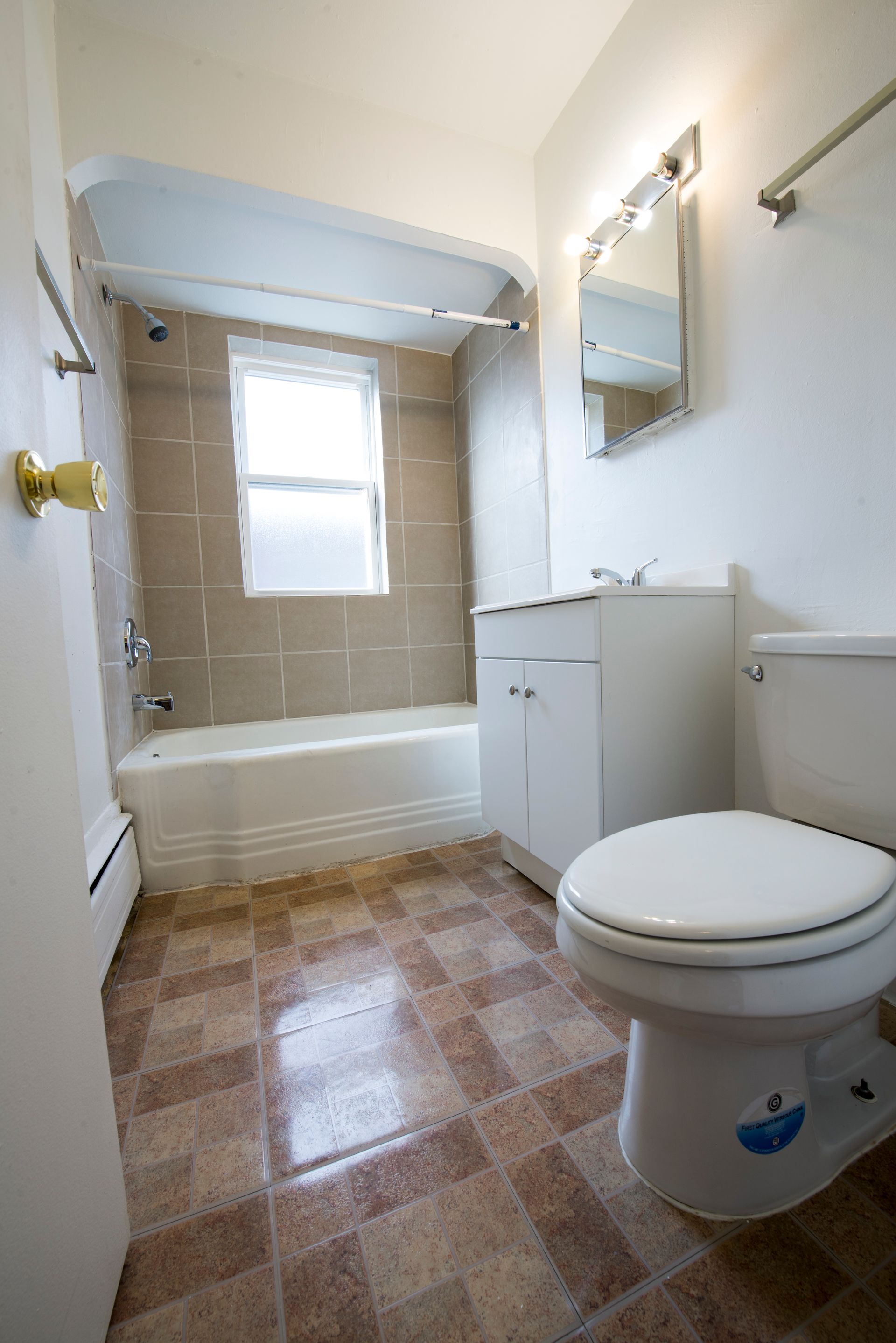 Bathroom with tan tiled walls, white tub, toilet, and vanity, and patterned floor.