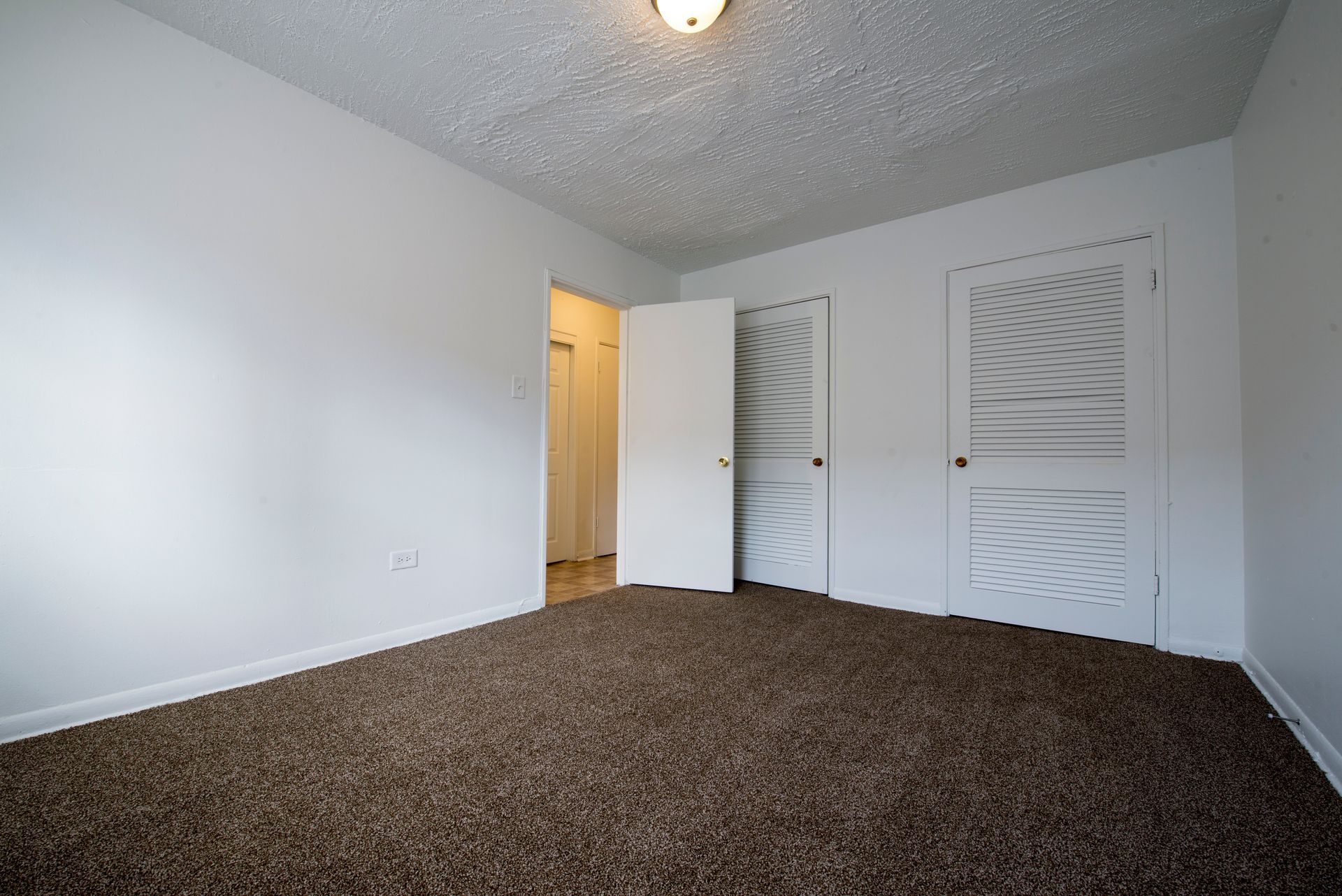 Empty room with white walls, brown carpet, textured ceiling, and closet doors.