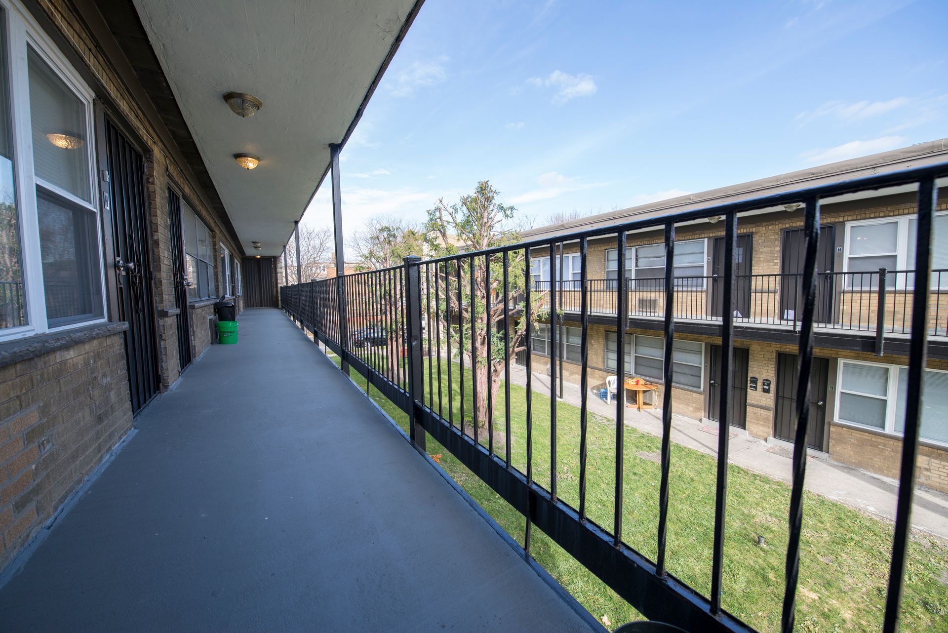 Exterior apartment walkway with black railing, overlooking grassy courtyard. Brick building, blue sky.