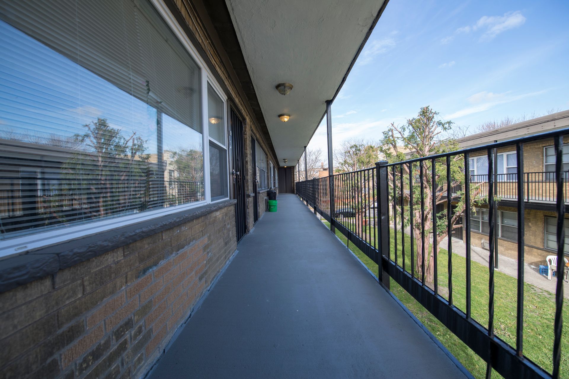 Apartment building exterior; long outdoor hallway with railing, windows, brick wall, and view of trees.