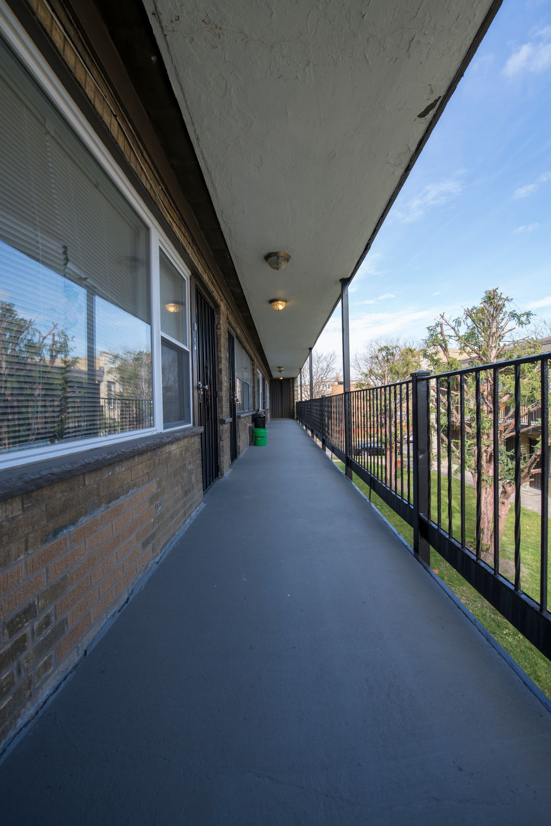 Narrow outdoor hallway of an apartment building, with windows on one side and a railing overlooking green space on the other.