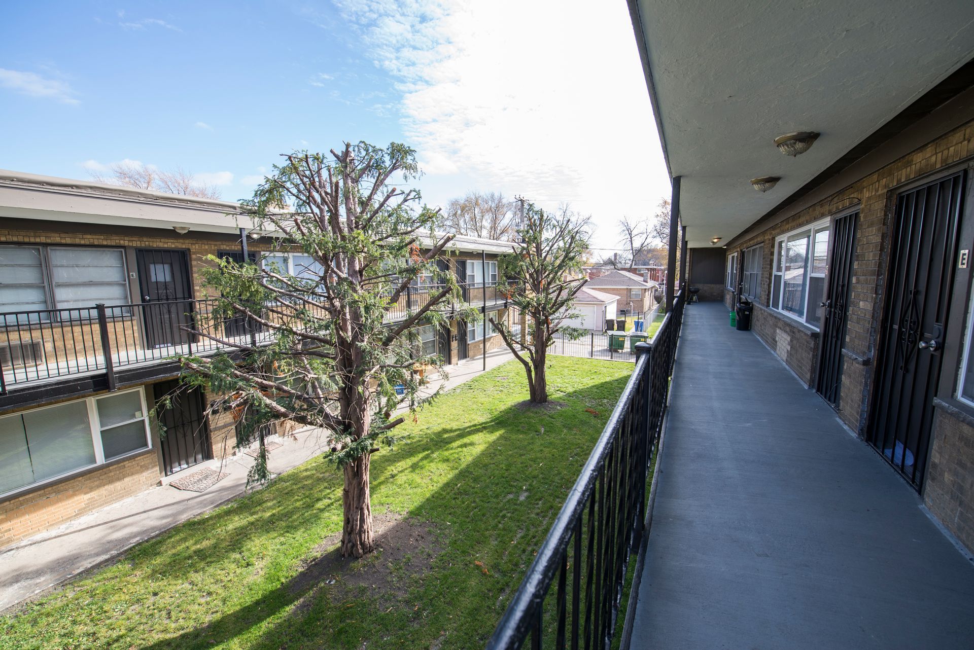 Two-story apartment building with exterior walkway, grassy area, and trees. Blue sky.