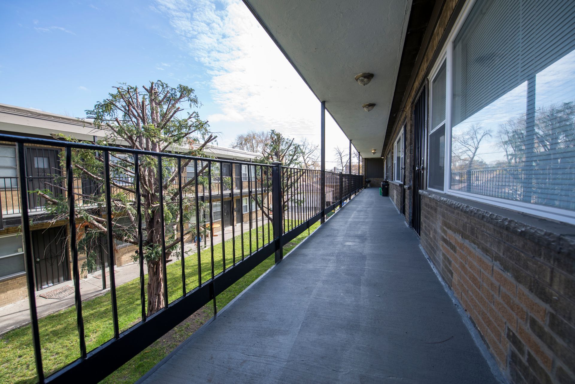 Exterior apartment walkway with black railing, windows, and brick wall.