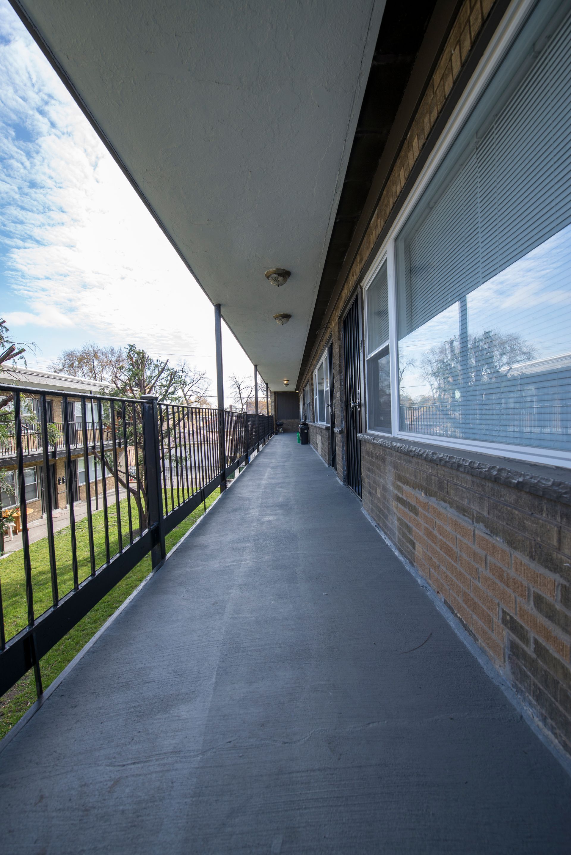 Exterior apartment hallway with a long, gray walkway, brick wall, and a black railing.