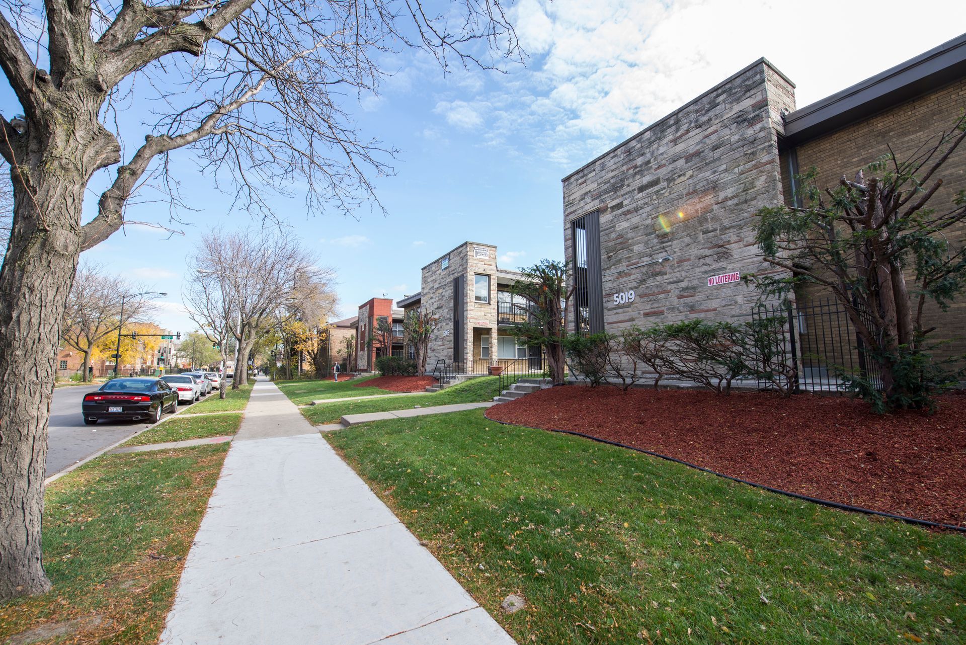 Sidewalk along brick apartment buildings with cars parked on street and trees.
