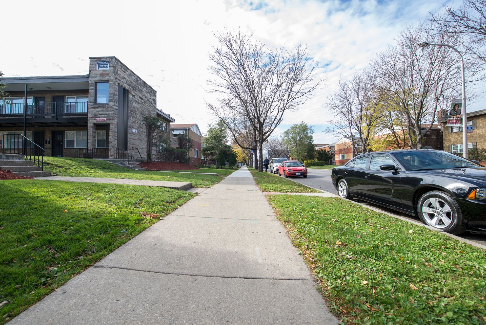 Sidewalk lined with trees and parked cars, two-story brick buildings, and grassy lawns.