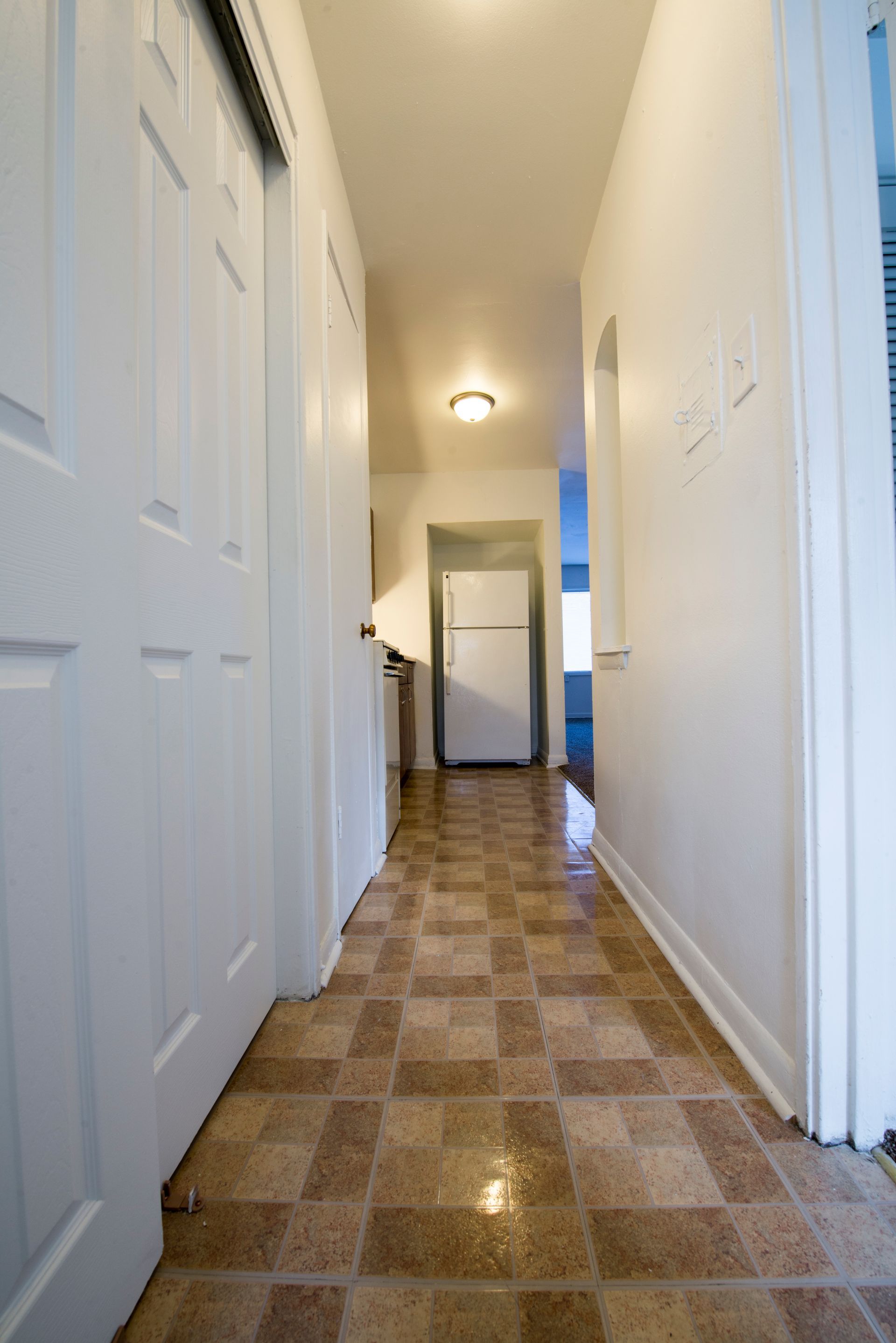 Narrow hallway with tiled floor, white doors, and a refrigerator at the end.