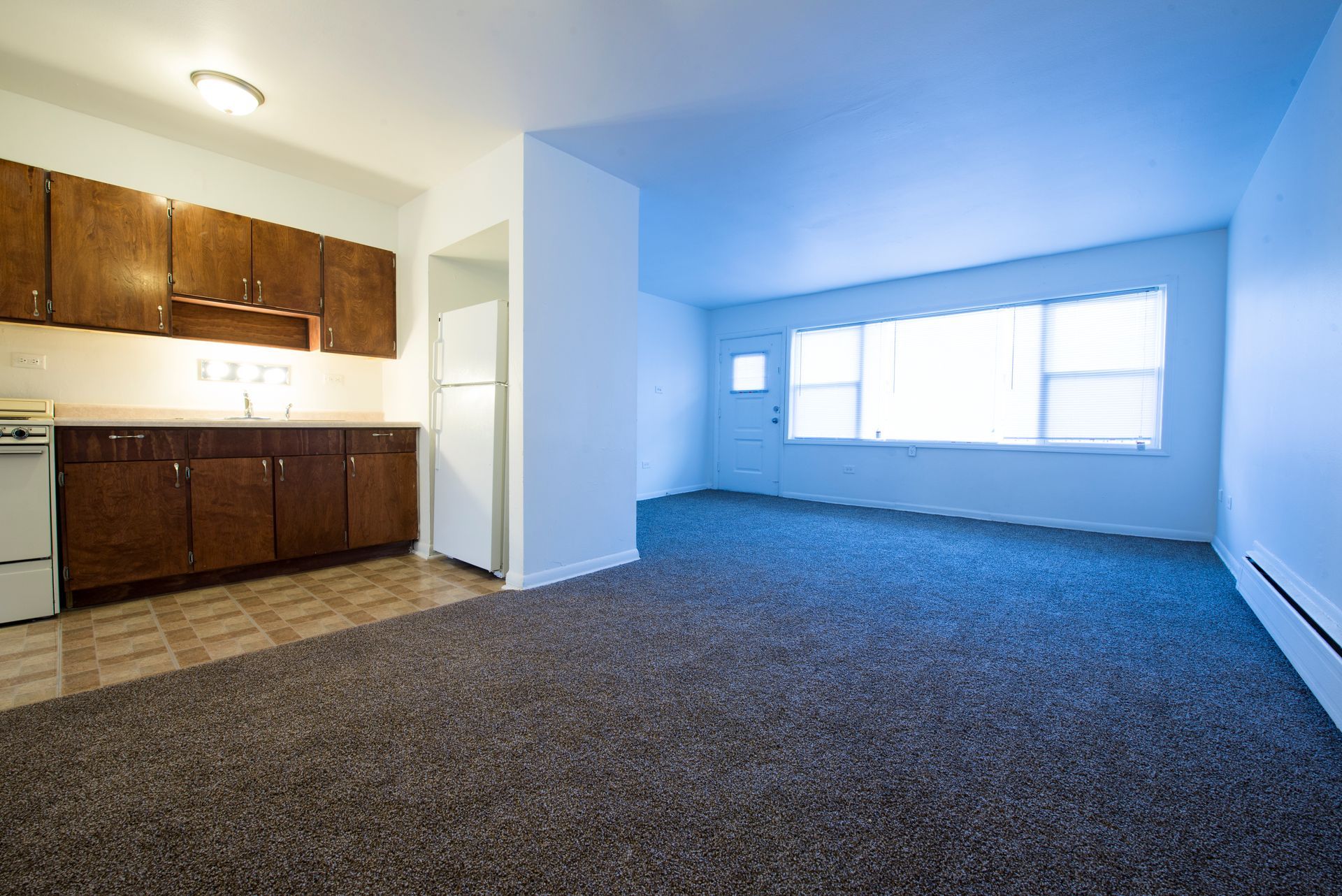 Apartment interior with kitchen and living area, brown cabinets, white appliances, and gray carpet.