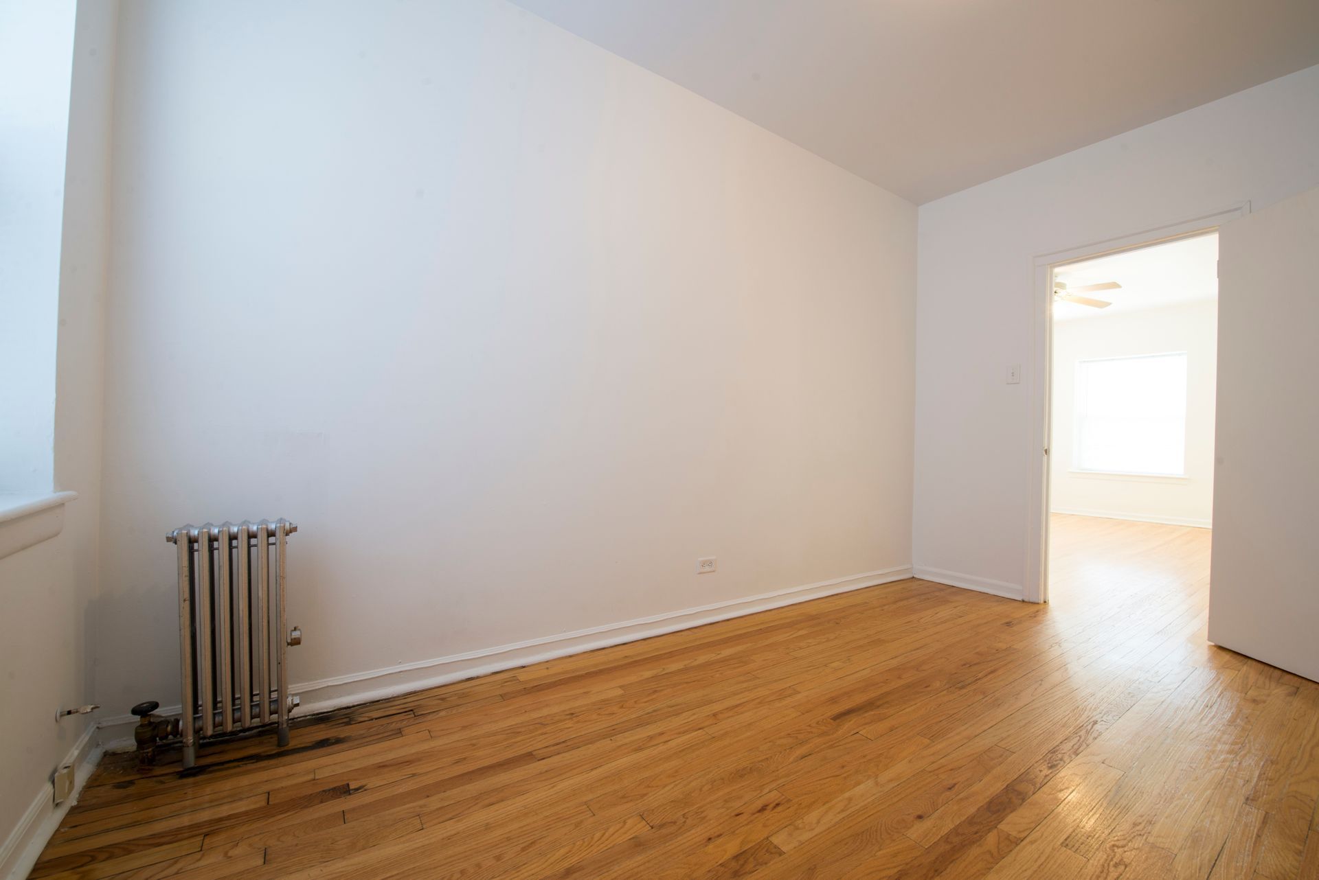 Empty room with hardwood floors, white walls, radiator, and doorway to another room.