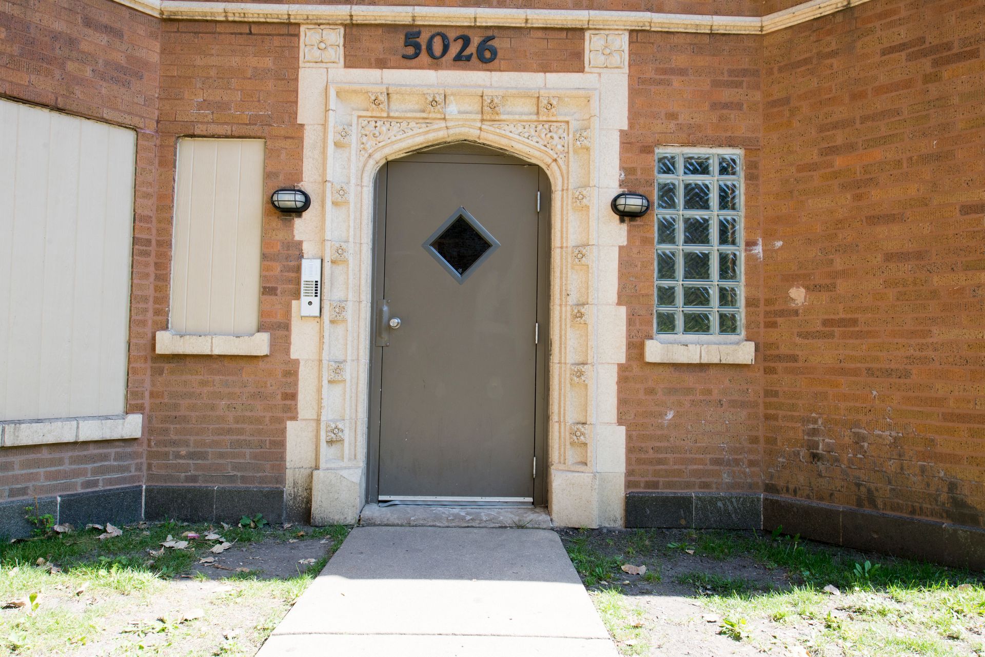 Brick building entrance with ornate stone door frame, gray door, small window, and address 5026.