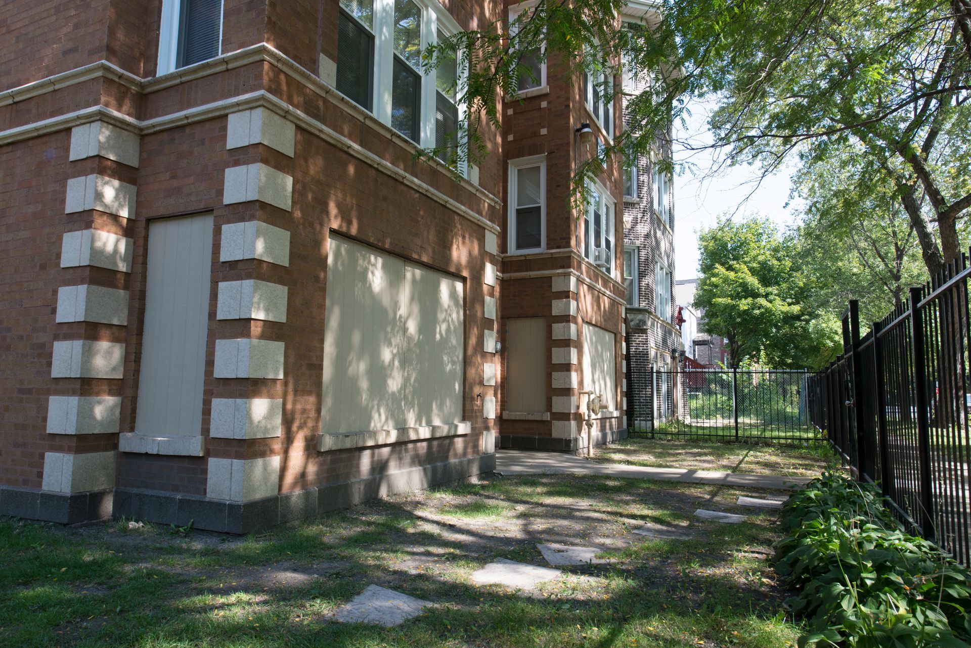 Brick building with boarded windows, beside a grassy area with a fence.
