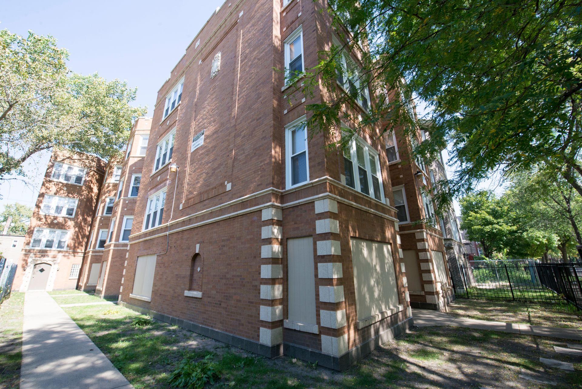 Brick apartment building with boarded-up windows, sidewalk, and trees.
