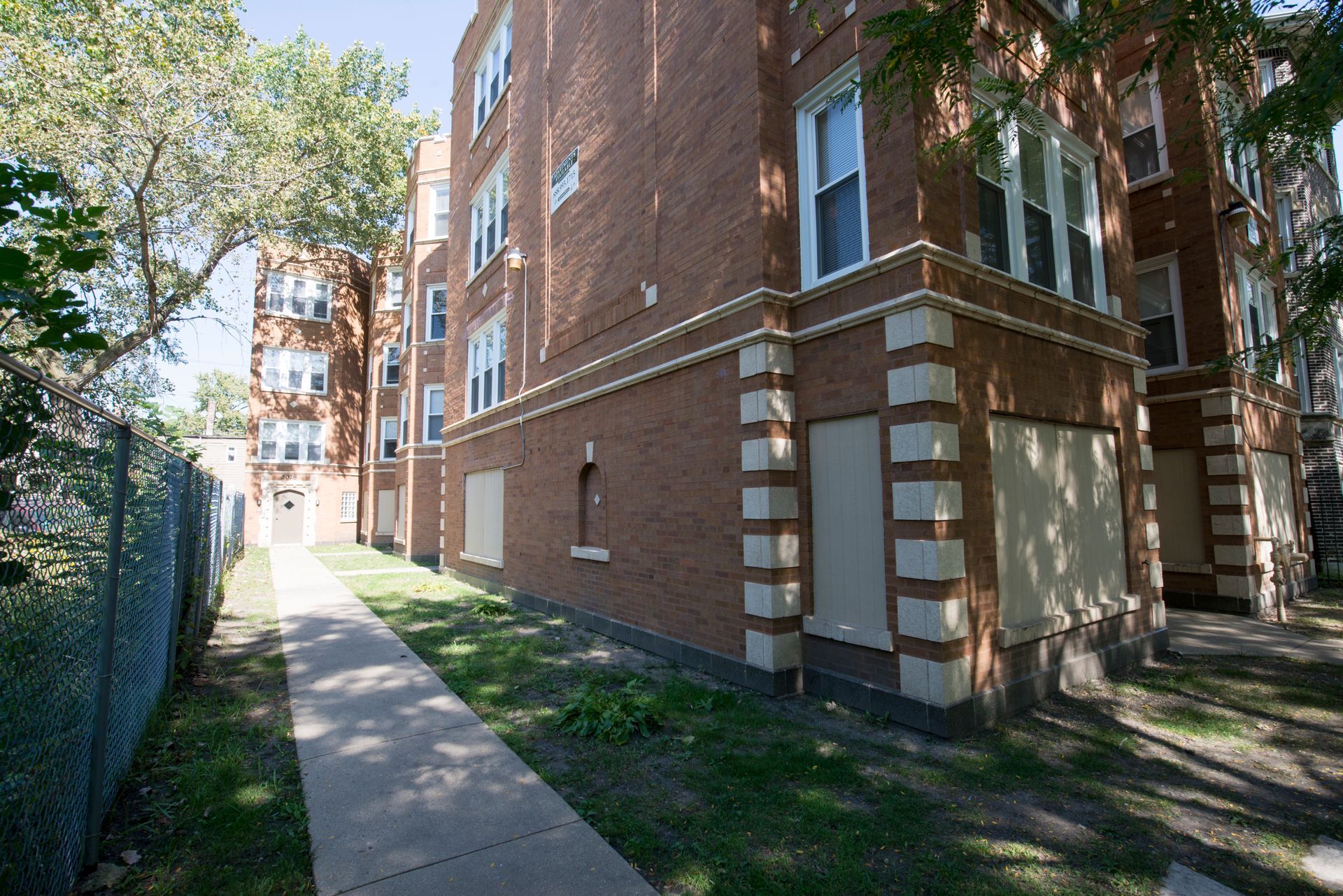 Brick apartment building with boarded-up windows, pathway, and chain-link fence.