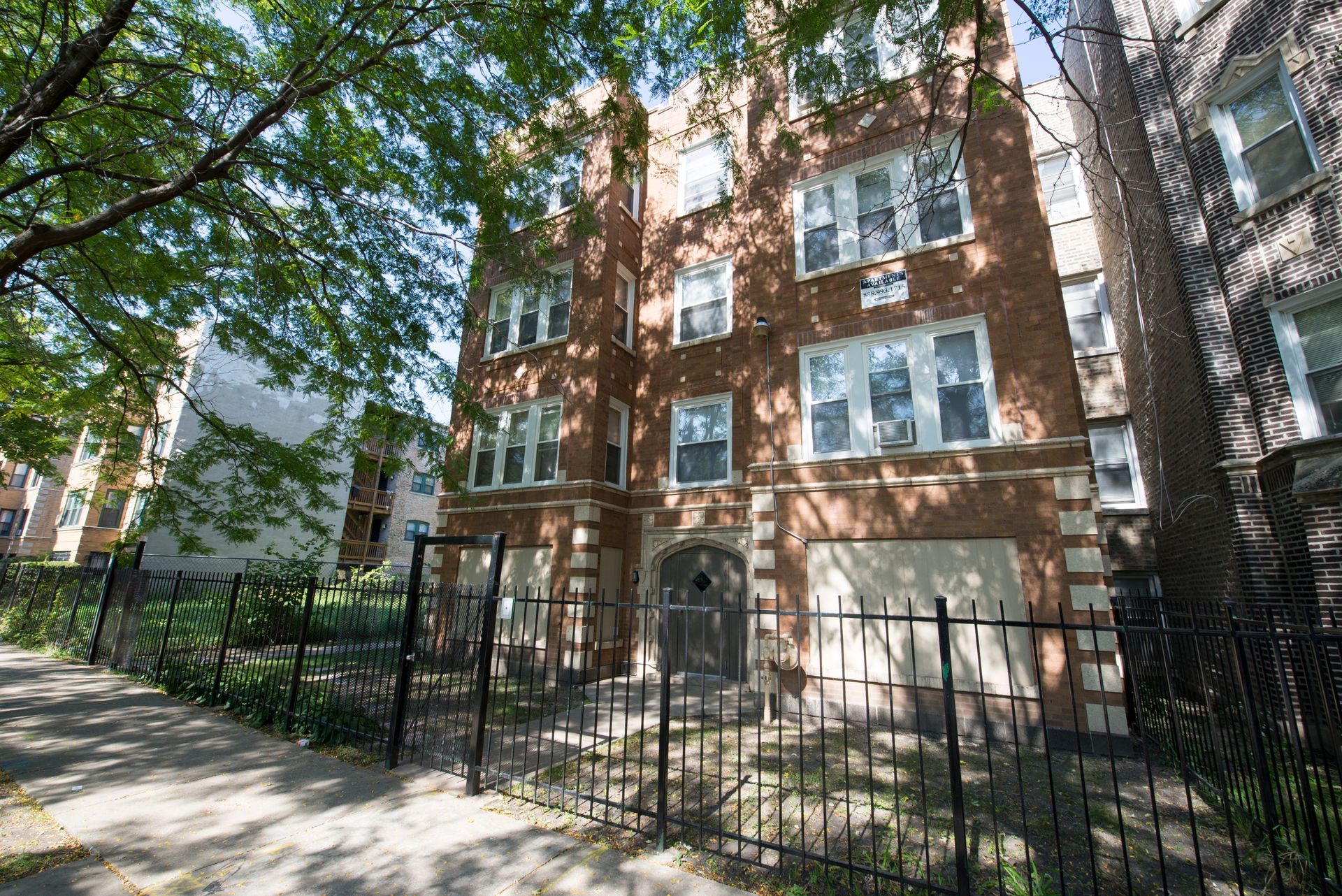 Brick apartment building with boarded-up windows behind a black metal fence on a city street.