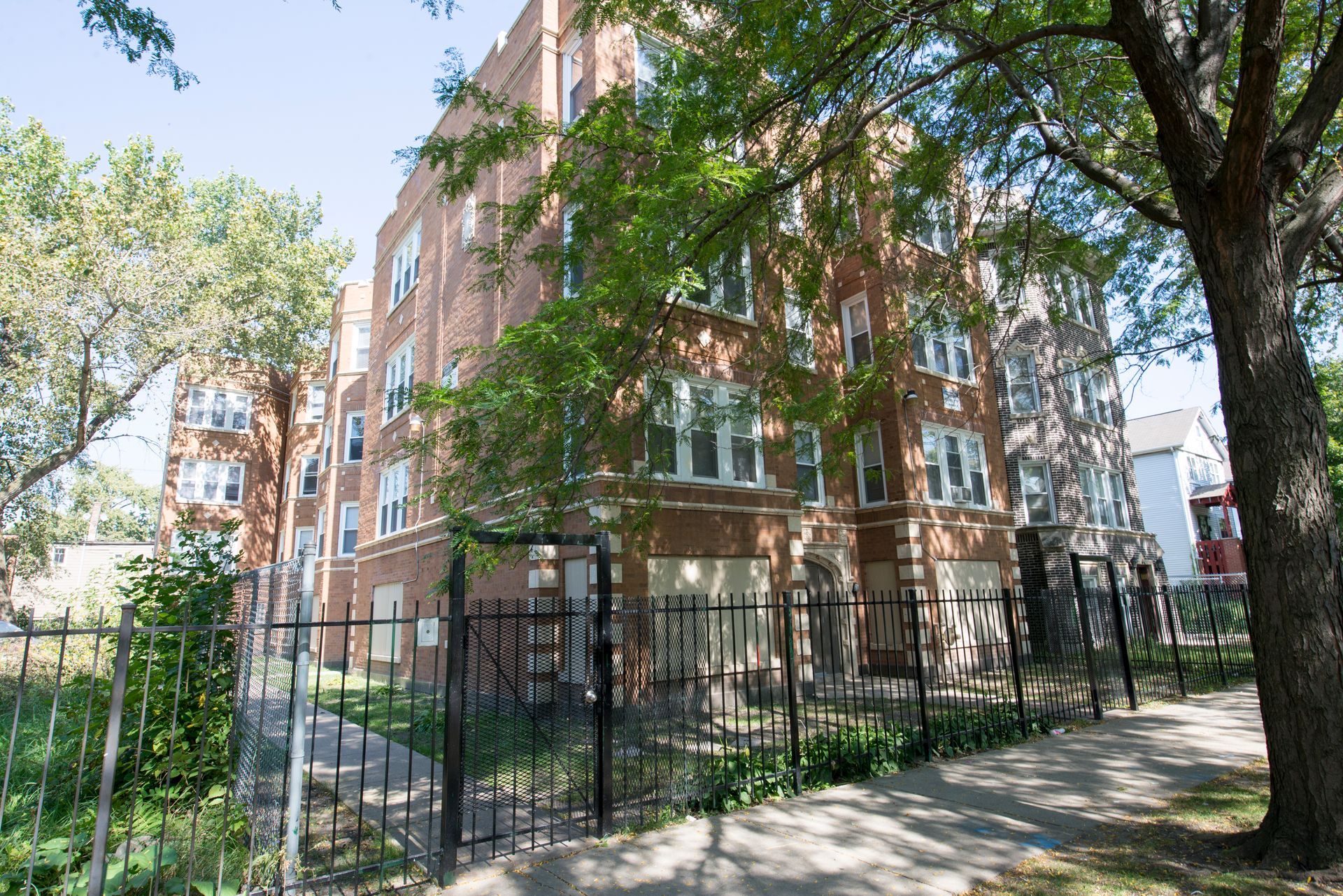 Brick apartment building with black fence and trees on a sunny day.