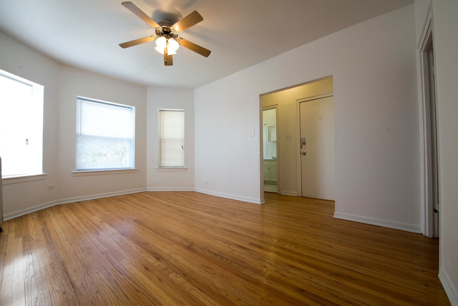 Empty room with hardwood floors, white walls, and a ceiling fan. Windows are on the left side, and a doorway leads to a bathroom.