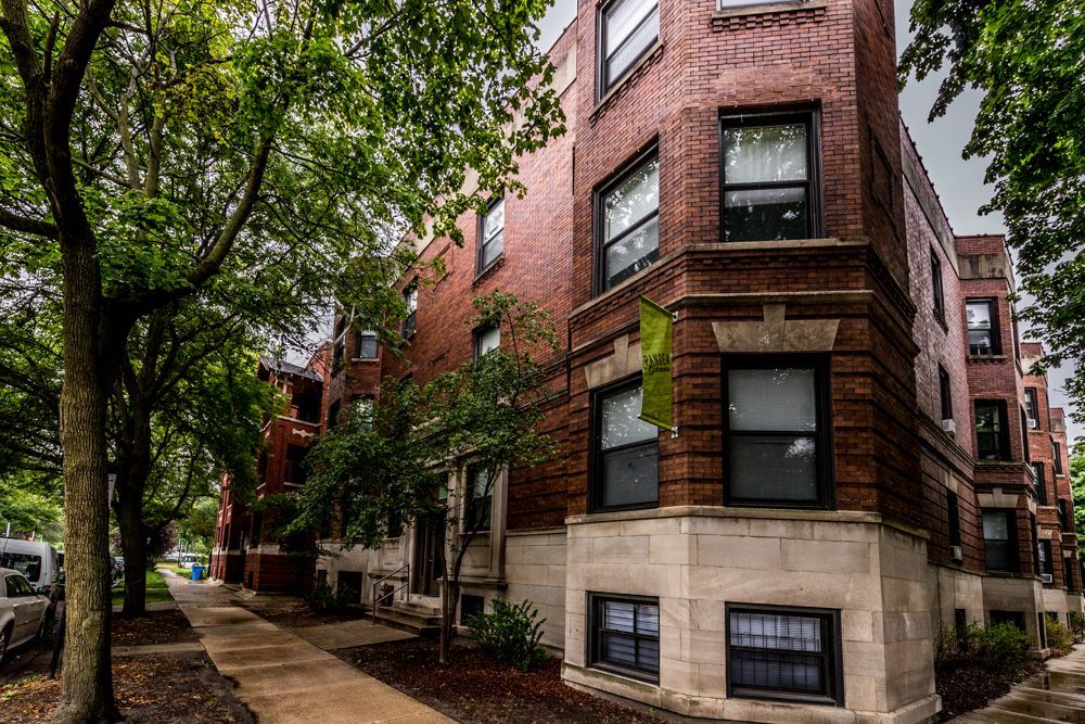 Brick apartment building with dark windows and a stone base; trees line the sidewalk.
