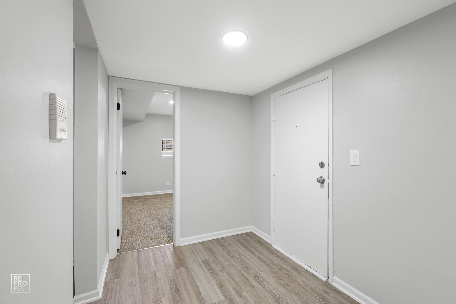Narrow hallway with gray walls, white door and trim, light wood-look flooring. Doorway to another room visible.