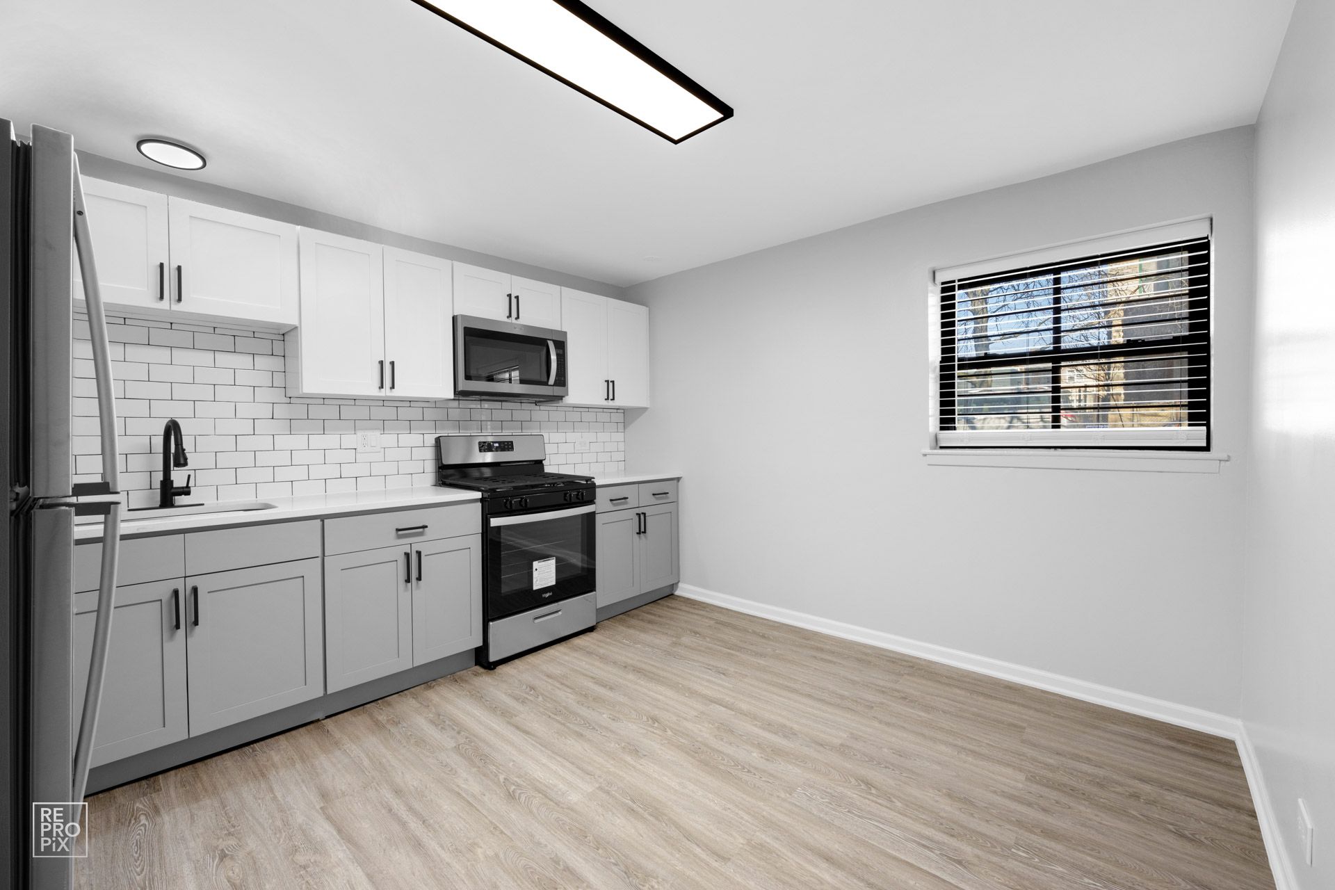 Kitchen with white and gray cabinets, stainless steel appliances, and wood-look flooring. Window with blinds.
