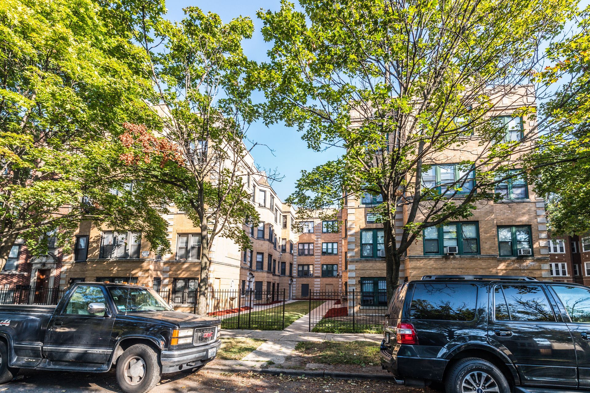 Apartment building exterior, trees line the street, cars parked in front.