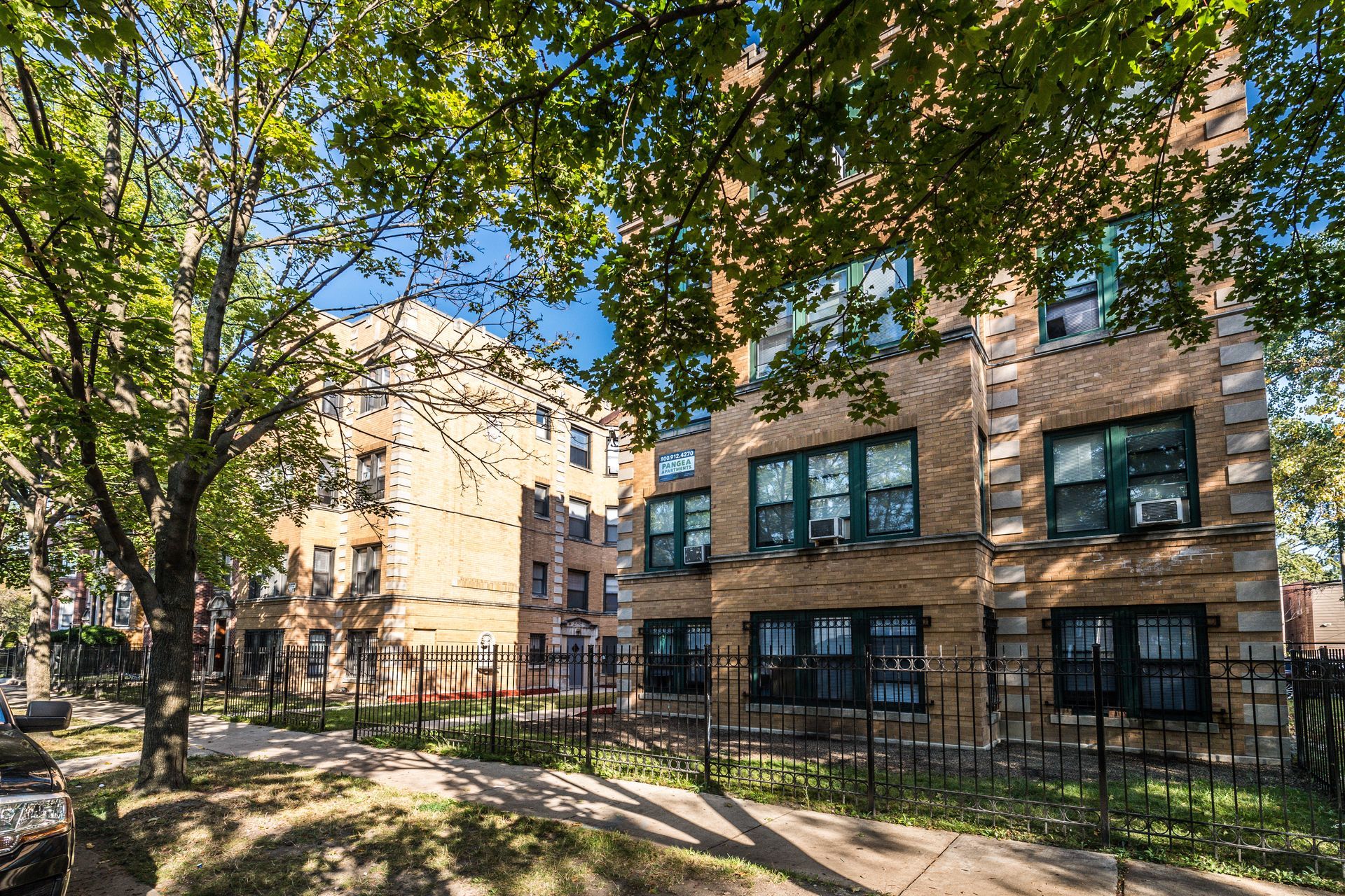Apartment buildings with brick facades, trees casting shadows on the sidewalk, and a wrought-iron fence.
