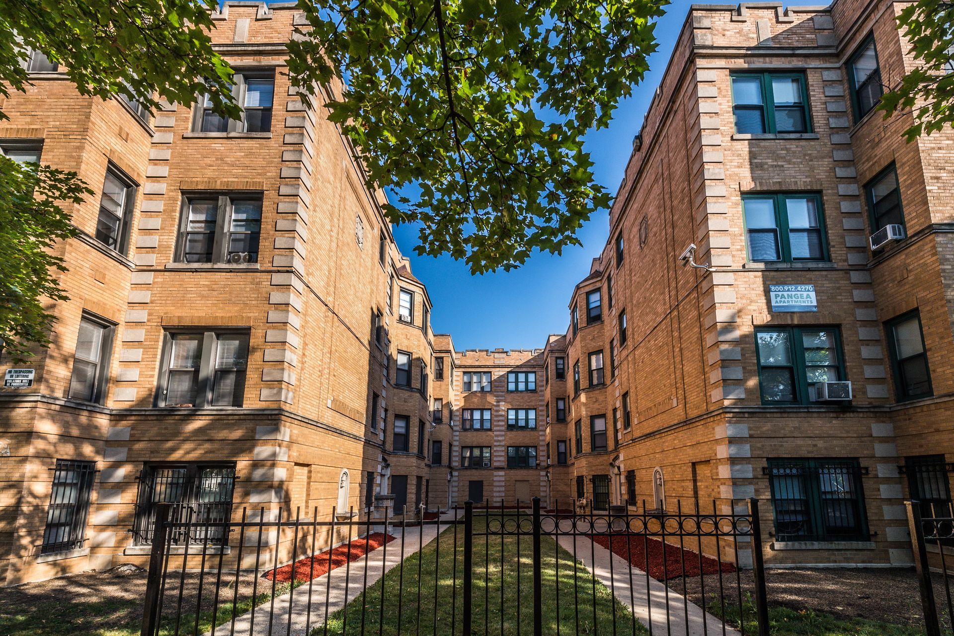 Brick apartment buildings form a courtyard, visible blue sky, framed by trees and a black fence.