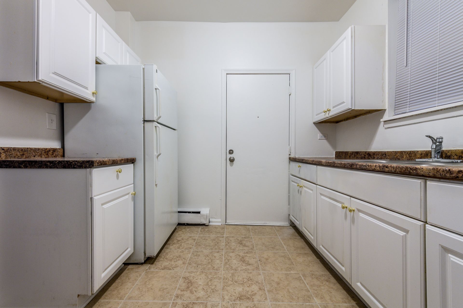 White kitchen with cabinets, refrigerator, and door. Brown countertops and tan floor tiles.