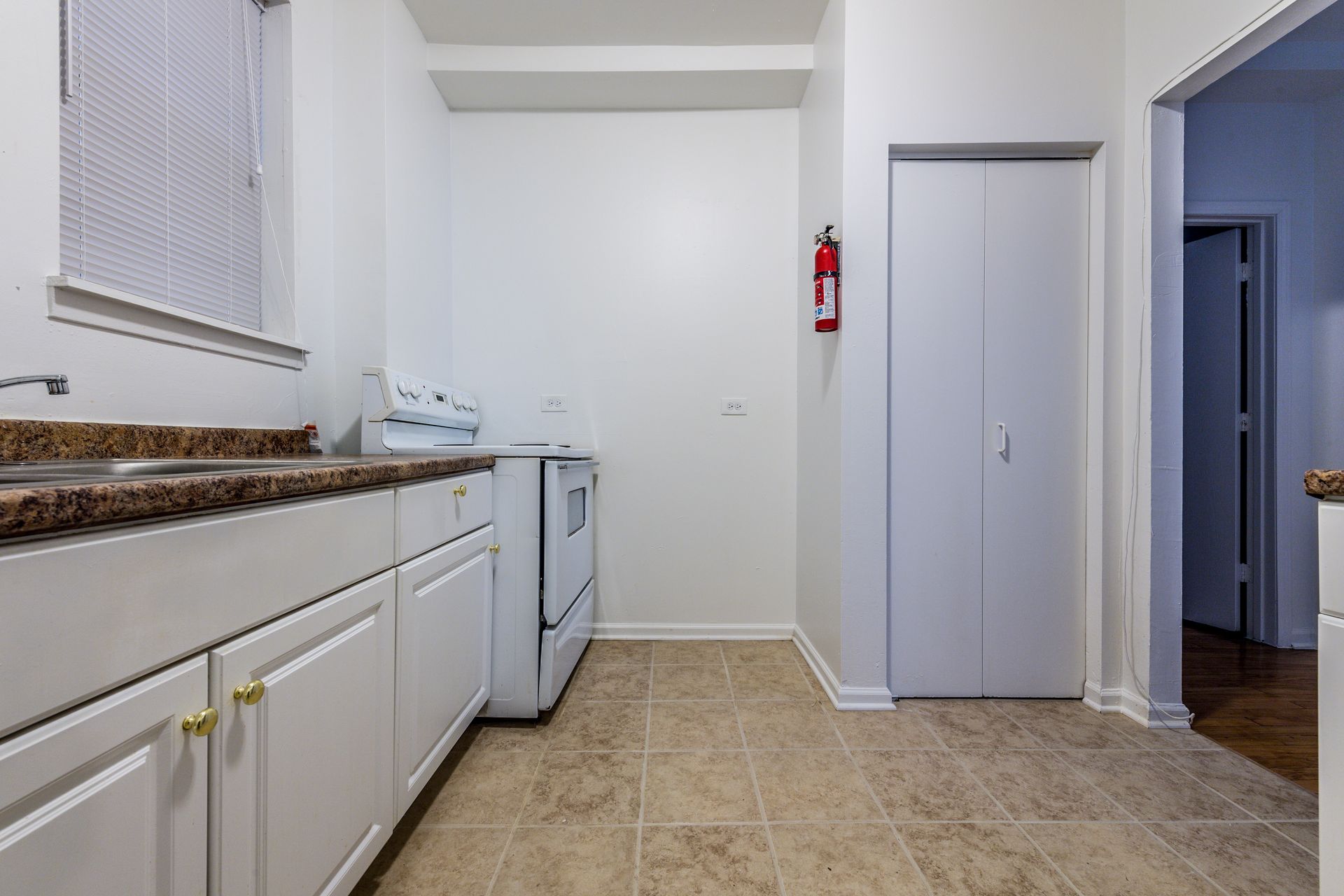 Small kitchen with white cabinets, stove, and door. Brown countertop, beige floor, and fire extinguisher.