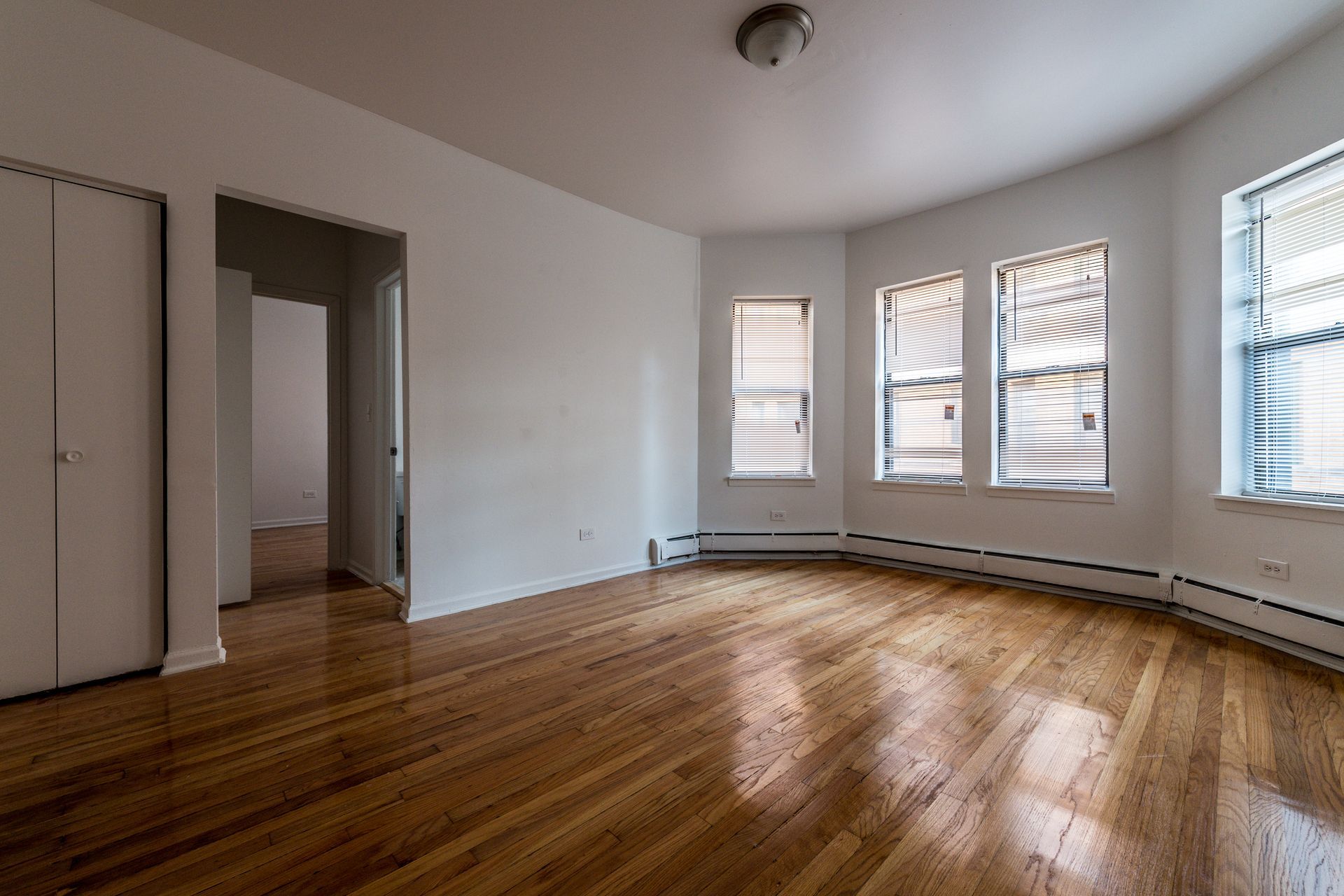 Empty room with hardwood floors, white walls, and three windows with blinds.