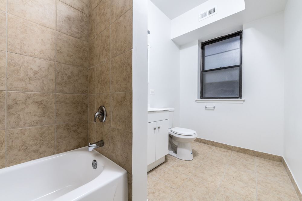 Bathroom with a bathtub, toilet, and window. Beige tile and flooring, white walls.