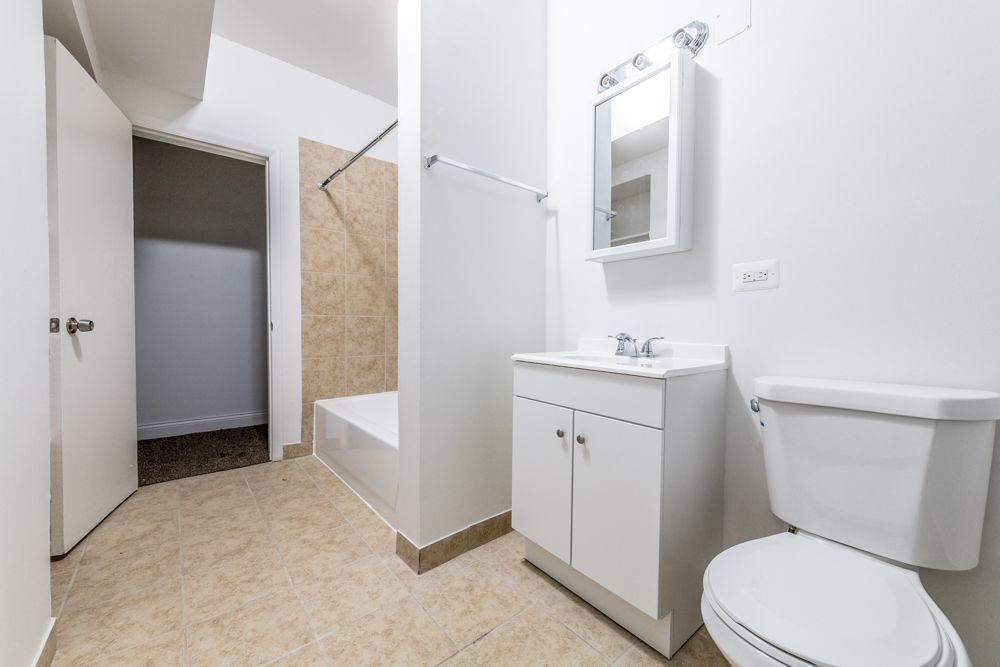 Bathroom with white walls, tan floor and shower tile, white vanity and toilet. Open doorway to closet.