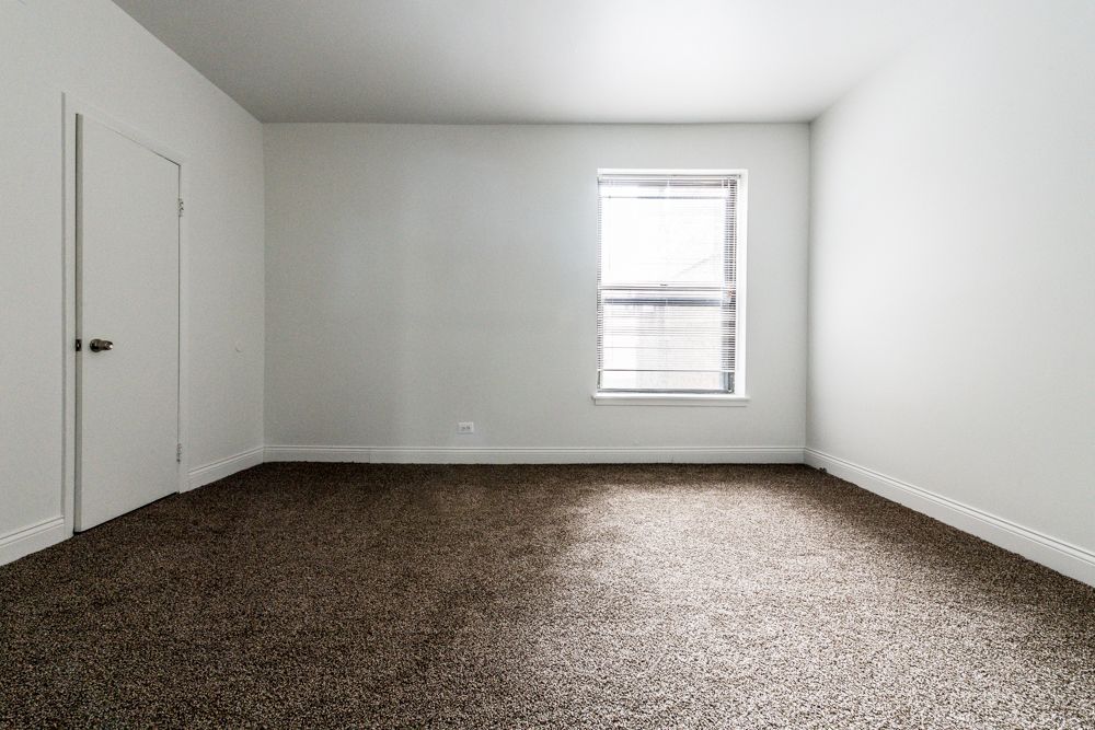 Empty room with white walls, a closed door, window with blinds, and brown carpet.
