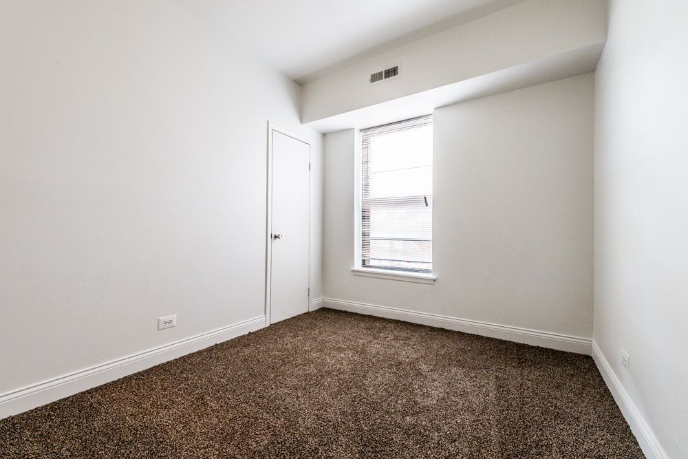 Empty room with brown carpet, white walls, small door, window with blinds.