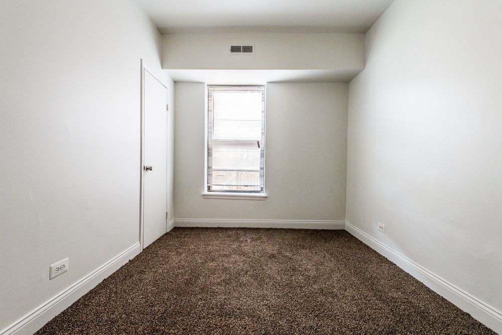 Empty room with carpet, a window, and a closed door. White walls, beige carpet, and a window with blinds.