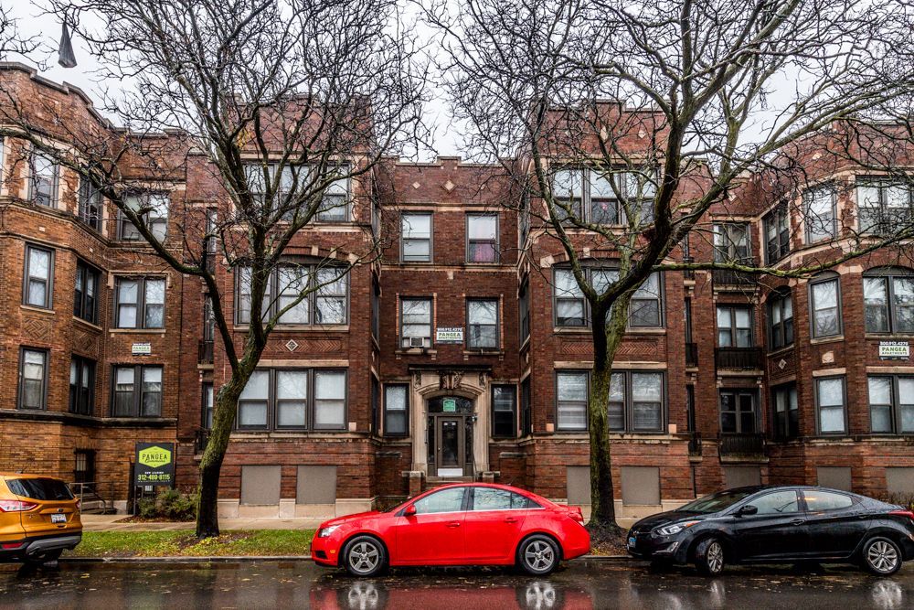 Brick apartment building with trees, parked cars, and cloudy sky.