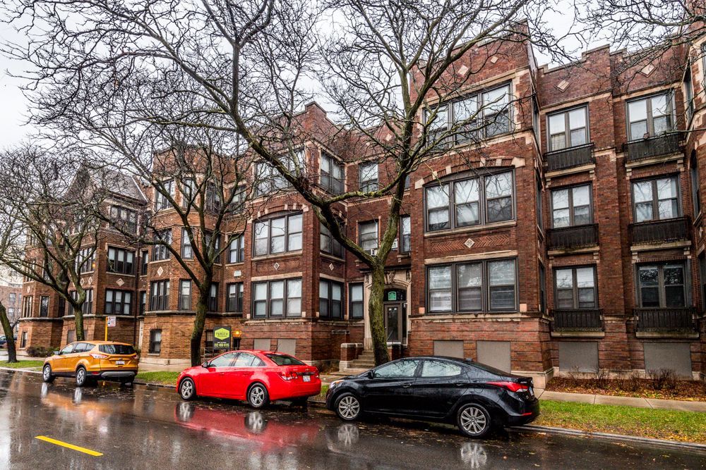 Apartment buildings along a wet street with parked cars and bare trees on a cloudy day.