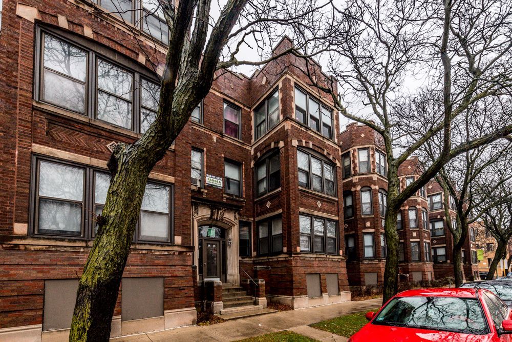 Red brick apartment building with multiple windows. A red car is parked on the street. Trees with bare branches.