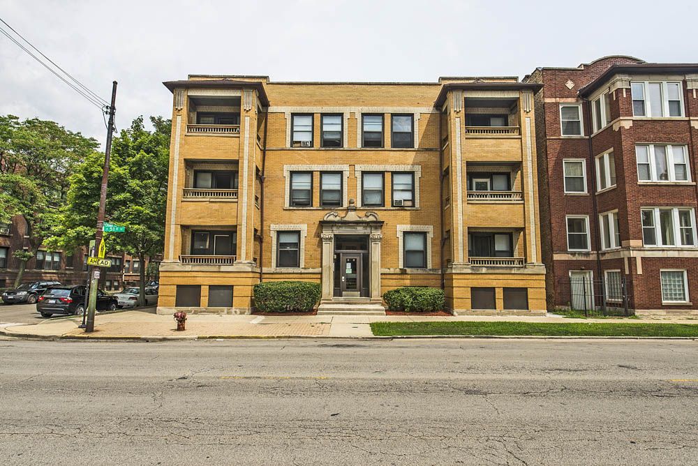 Yellow brick apartment building with architectural details; sidewalk and street in front.