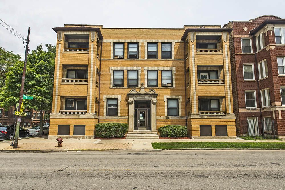 Yellow brick apartment building with balconies, sidewalk, street, and adjacent building.