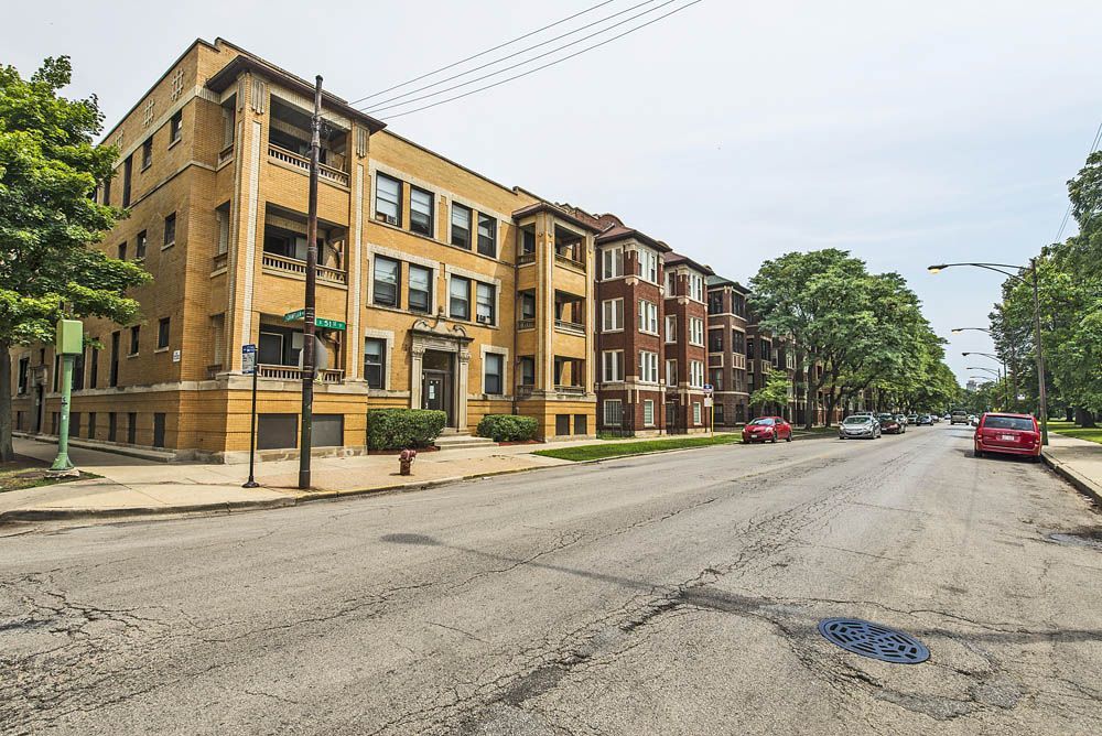 Apartment buildings line a cracked street with parked cars and trees under a cloudy sky.
