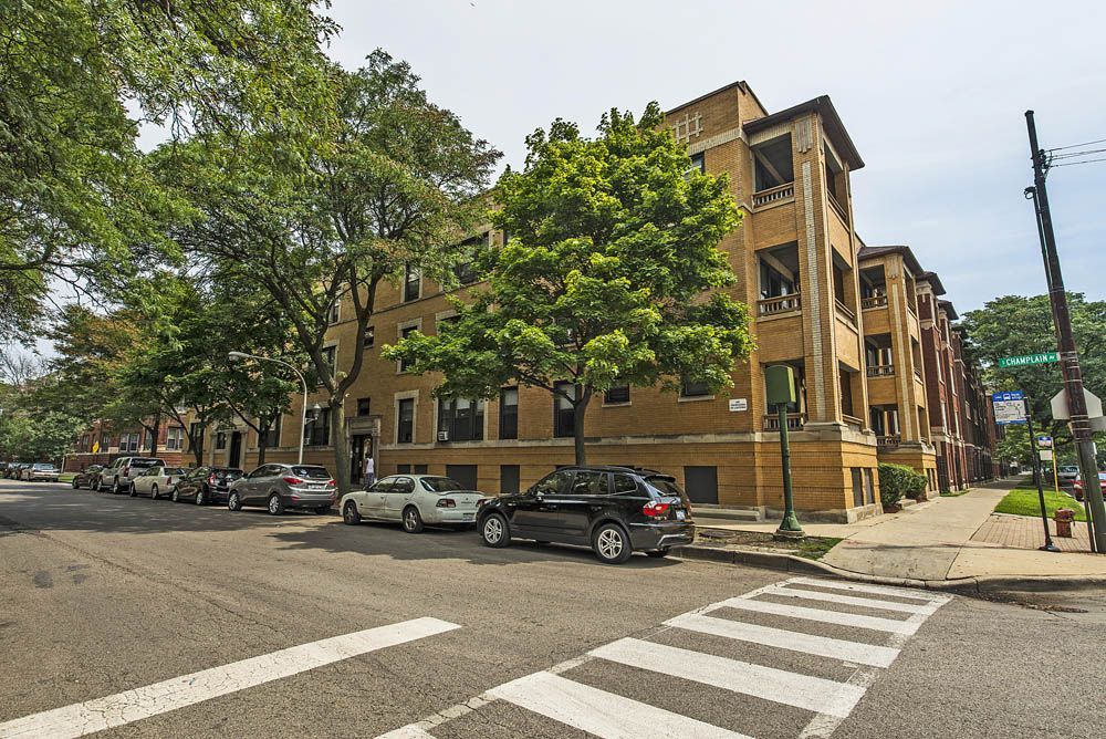 Apartment building on a tree-lined street with parked cars. Crosswalk and street sign visible.