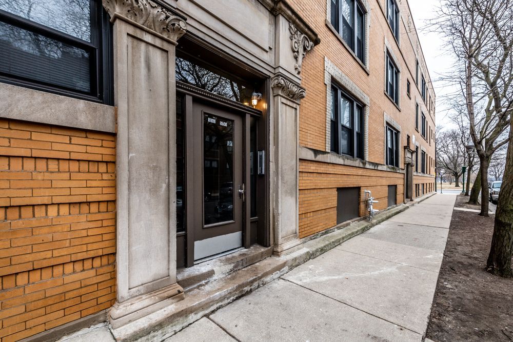 Building entrance with brick and stone facade, sidewalk, and trees.